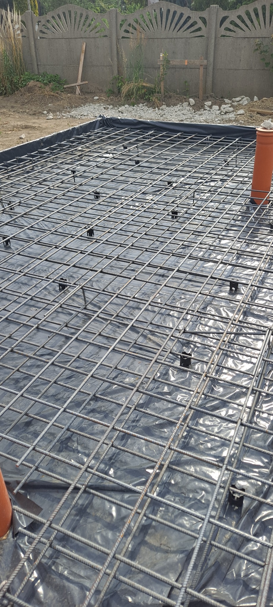 Zbrojenie fundamentów z siatki stalowej na czarnej folii izolacyjnej, widoczne plastikowe dystanse i rura kanalizacyjna. W tle betonowy płot i fragment terenu.