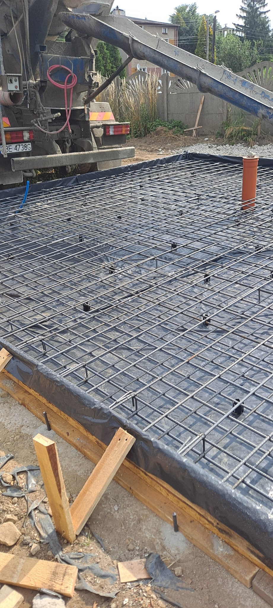 Zbrojenie fundamentu z siatki stalowej, przygotowane do zalewania betonem z betoniarki widocznej w tle, z pomarańczową rurą kanalizacyjną wystającą z przygotowanej powierzchni.