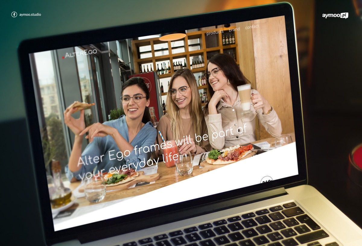 Ekran laptopa wyświetlający stronę internetową AlterEgo (oprawy okularowe) z trzema uśmiechniętymi kobietami w okularach, siedzącymi przy stole w kawiarni z pizzą i napojami.