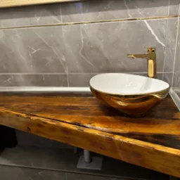 Umywalka nablatowa w kolorze białym ze złotym rantem i złota bateria na drewnianym blacie o nieregularnej krawędzi w łazience wykończonej szarymi płytkami imitującymi marmur.