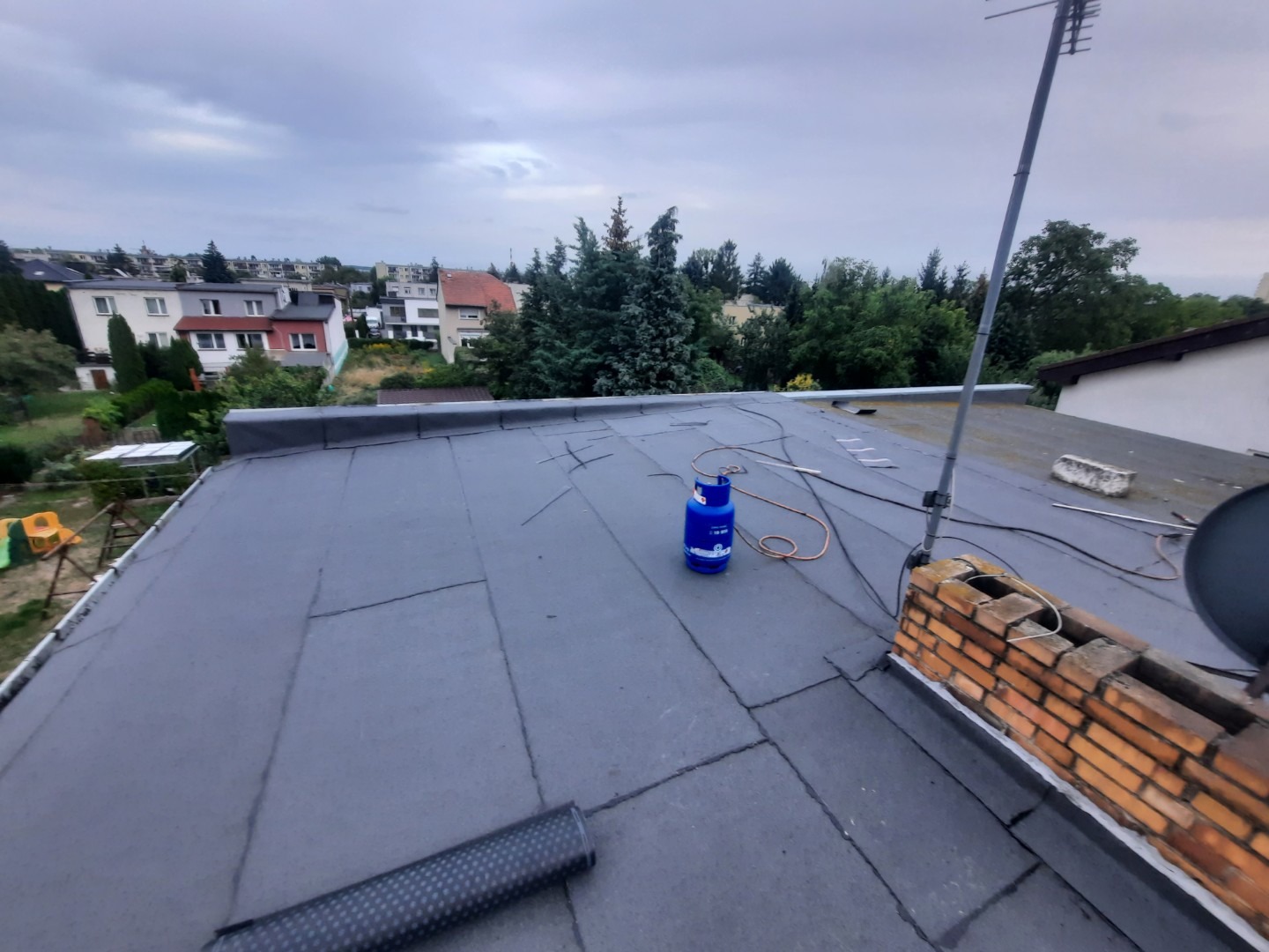 Płaski dach pokryty papą, z widocznym kominem, anteną, butlą gazową i zwiniętym pasem papy w rogu; w tle zabudowa jednorodzinna i zieleń.