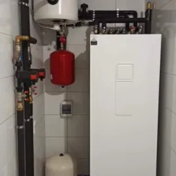 Zainstalowany system grzewczy z białym zbiornikiem, czerwonym naczyniem przeponowym i czarnymi rurami w pomieszczeniu wyłożonym białymi płytkami.