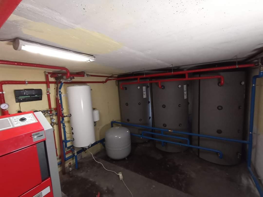 Kotłownia z trzema szarymi zbiornikami akumulacyjnymi ciepła, białym zasobnikiem CWU, czerwonym piecem, oraz instalacją z rur w kolorze czerwonym i niebieskim, w piwnicy z betonową podłogą i sufitem.