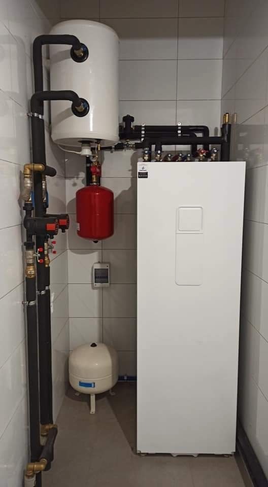 Zainstalowany system grzewczy z białym zbiornikiem, czerwonym naczyniem przeponowym i czarnymi rurami w pomieszczeniu wyłożonym białymi płytkami.