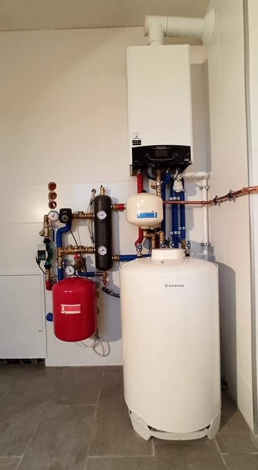 Zestaw kotła gazowego Ariston z zasobnikiem ciepłej wody i osprzętem hydraulicznym na tle jasnej ściany i szarej podłogi.