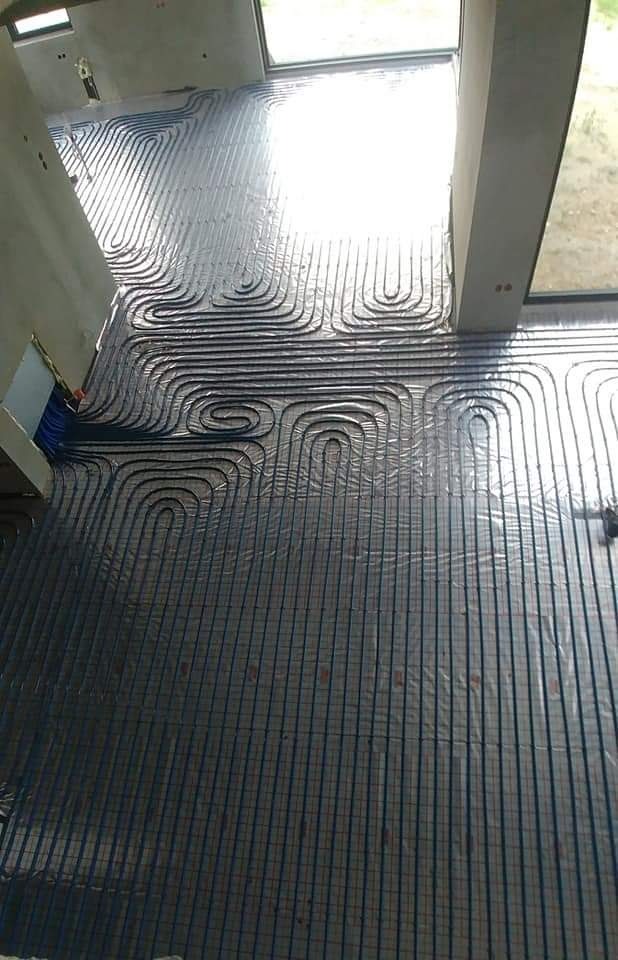 Widok z góry na rozłożone rury ogrzewania podłogowego na folii izolacyjnej w surowym stanie, przed wylaniem posadzki, z widocznym fragmentem niebieskich rur przyłączeniowych.