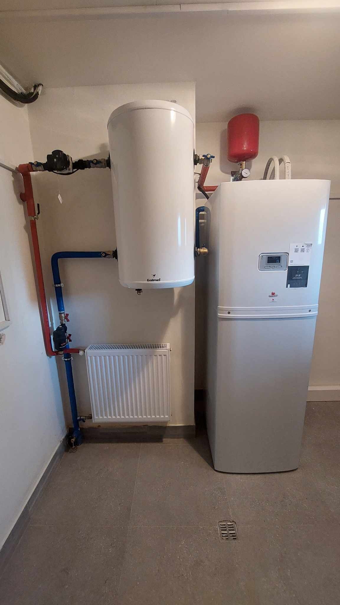 Biały zbiornik buforowy Galmet zawieszony nad grzejnikiem, obok biała jednostka pompy ciepła z czerwonym zbiornikiem wyrównawczym, instalacja w kolorze czerwonym i niebieskim, szara podłoga...