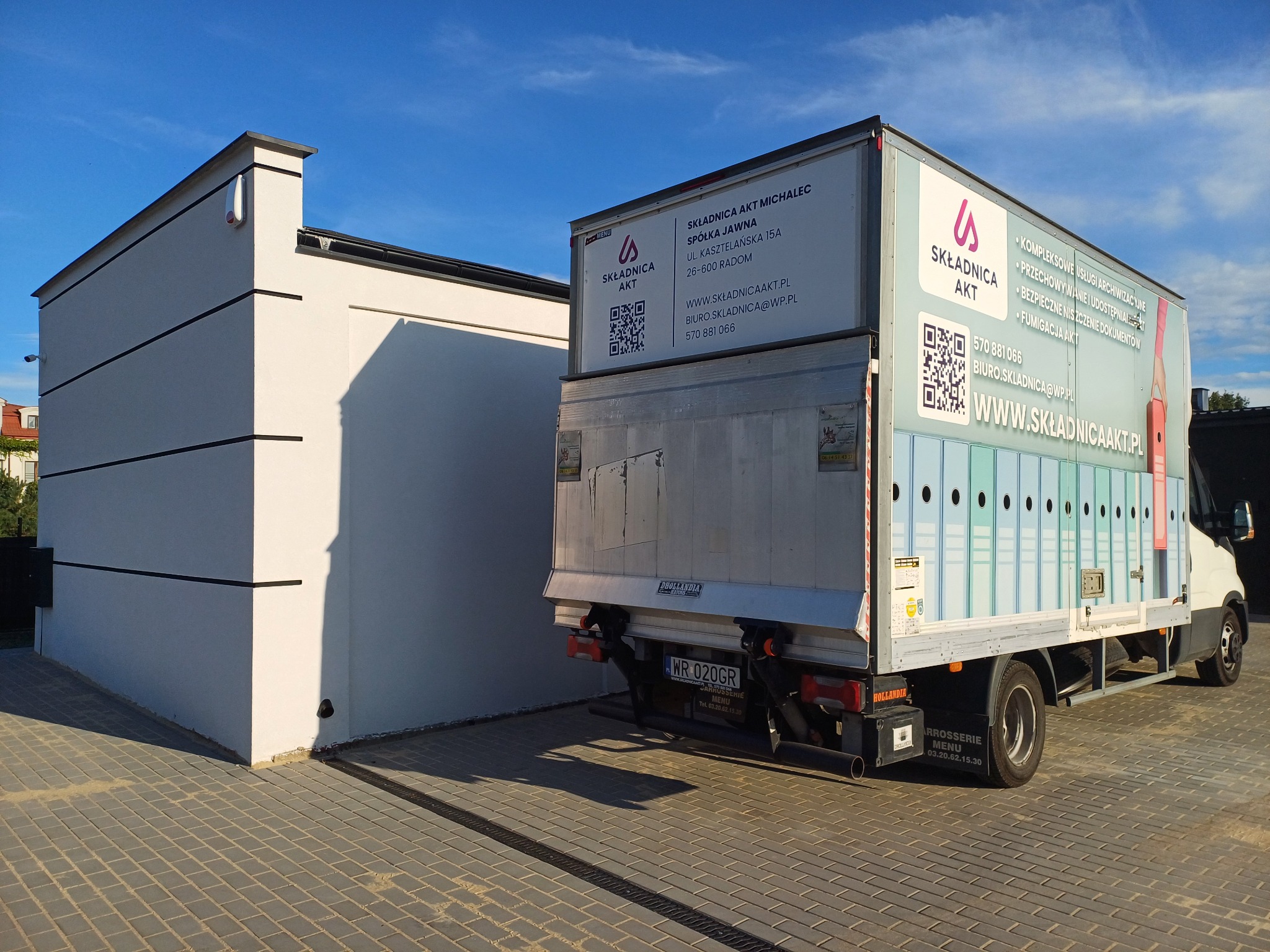 Samochód dostawczy firmy Składnica Akt z otwartymi drzwiami, zaparkowany na brukowanej powierzchni przed nowoczesnym budynkiem o białej elewacji, reklama na samochodzie informuje o kompleksowych...