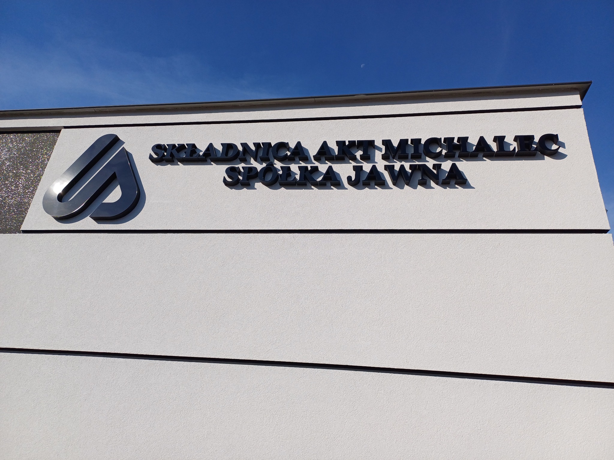 Elewacja budynku z logo i nazwą firmy: Składnica Akt Michalec Spółka Jawna, widoczne na tle błękitnego nieba.