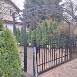 Czarna brama wjazdowa z metalowymi zdobieniami i łukiem, widoczna z zewnątrz posesji, obok siatka ogrodzeniowa i tuje.