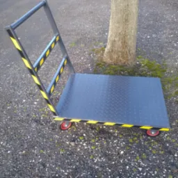 Wózek transportowy z metalową platformą o wzorze rombów, pomalowany na szaro i oznaczony żółto-czarnymi pasami ostrzegawczymi, stojący na żwirowej nawierzchni obok pnia drzewa.