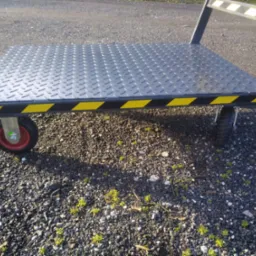 Metalowy wózek platformowy z ryflowaną powierzchnią i żółto-czarnymi pasami ostrzegawczymi, stojący na żwirowym podłożu z kiełkującą roślinnością.