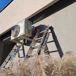 FAME INSTAL - Zewnętrzna jednostka klimatyzacji marki Kaisai zamontowana na ścianie budynku, widoczna z perspektywy niskiego kąta przez ozdobne trawy, drabina Krause oparta o ścianę, błękitne niebo w tle.