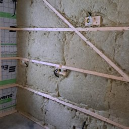 FAME INSTAL - Ściana w trakcie izolacji wełną mineralną, widoczne drewniane legary, puszki elektryczne i przewody, surowy beton na podłodze.