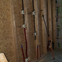 FAME INSTAL - Wewnętrzna instalacja elektryczna i rury w trakcie montażu w drewnianej konstrukcji ściany, widoczne kable w peszlach i puszki elektryczne, narzędzie podpierające konstrukcję, surowy stan wykończenia.