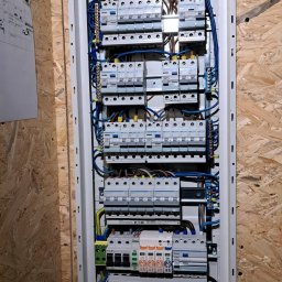 FAME INSTAL - Rozdzielnica elektryczna z bezpiecznikami, zamontowana na ścianie z płyty OSB, widoczne przewody w kolorach żółto-zielonym, niebieskim i brązowym, oraz biała obudowa rozdzielnicy.
