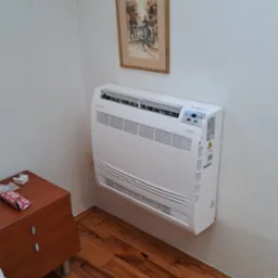 Biały klimatyzator podłogowy marki Rotenso Aneru, zamontowany w rogu pokoju na drewnianej podłodze, widoczny obok szafki nocnej i obrazka na ścianie.