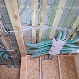 Instalacja rekuperacji i przewodów elektrycznych w surowym drewnianym wnętrzu, widoczne rury wentylacyjne w kolorze seledynowym i kable elektryczne przymocowane do drewnianej konstrukcji stropu.