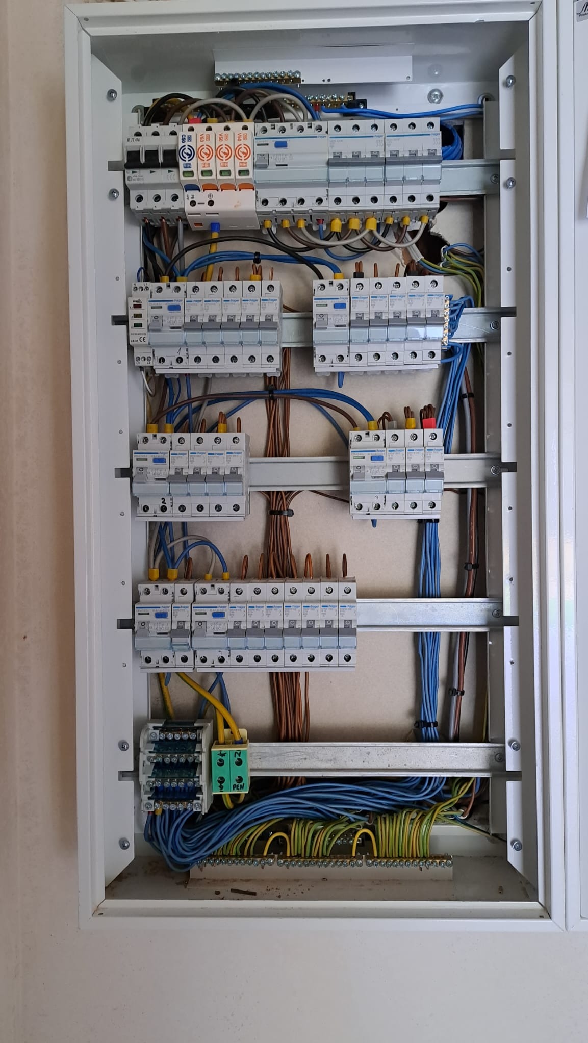 Otwarta skrzynka elektryczna z zainstalowanymi bezpiecznikami, przewodami w różnych kolorach i listwami zaciskowymi, widoczne elementy montażowe i oznaczenia na urządzeniach.