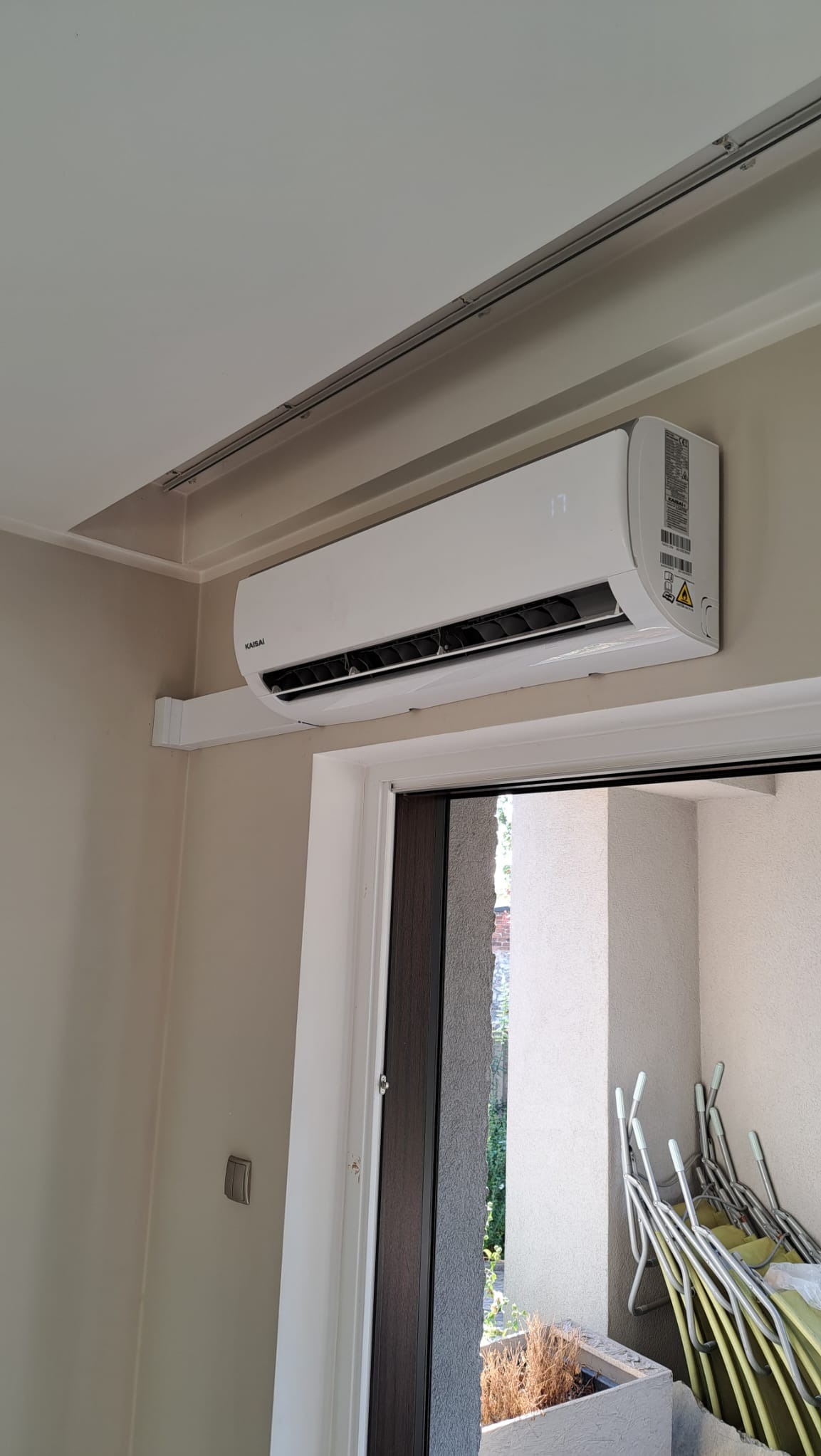 Biały klimatyzator Kaisai zamontowany na ścianie nad drzwiami balkonowymi, z widocznym wyświetlaczem wskazującym temperaturę 17 stopni Celsjusza, w tle widok na balkon z narzędziami ogrodniczymi.
