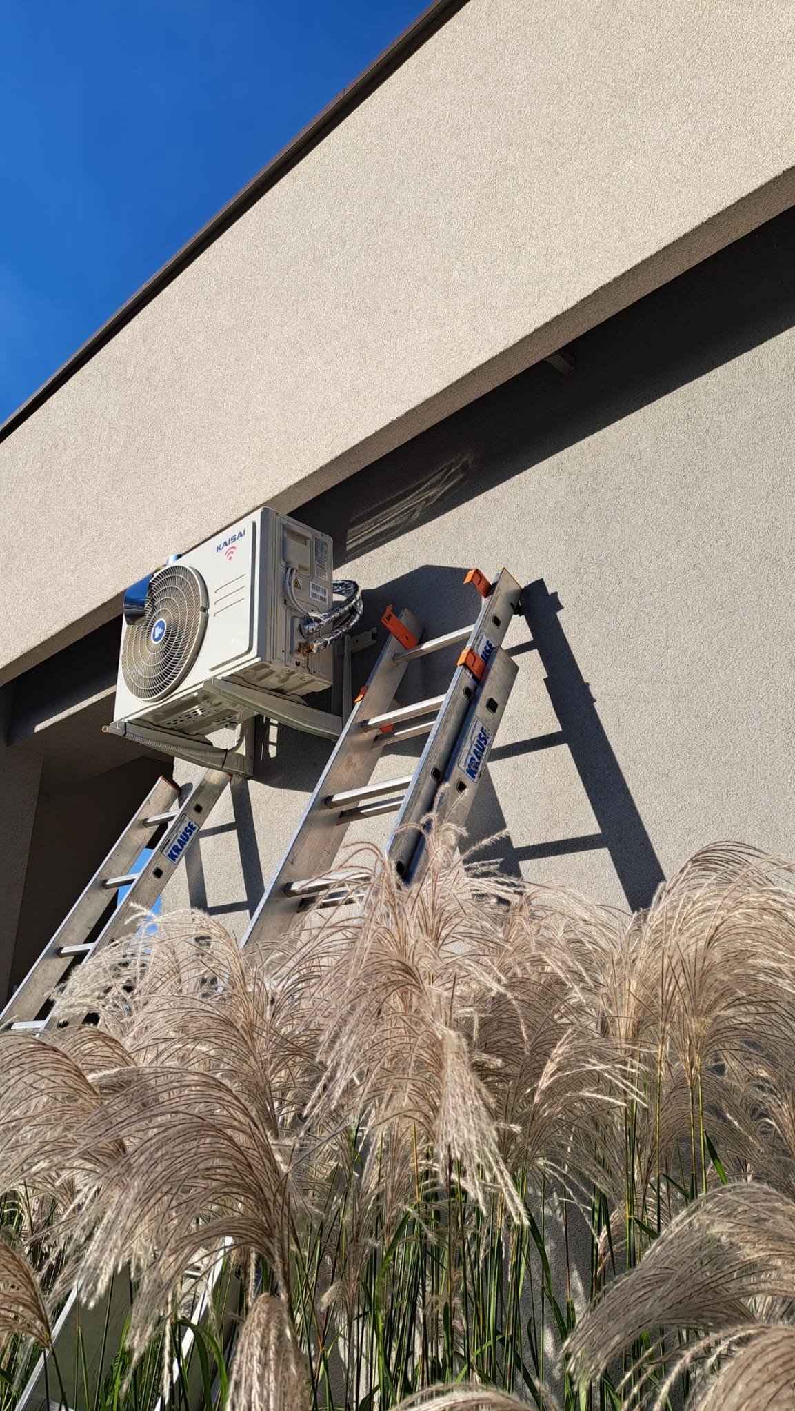 Zewnętrzna jednostka klimatyzacji marki Kaisai zamontowana na ścianie budynku, widoczna z perspektywy niskiego kąta przez ozdobne trawy, drabina Krause oparta o ścianę, błękitne niebo w tle.