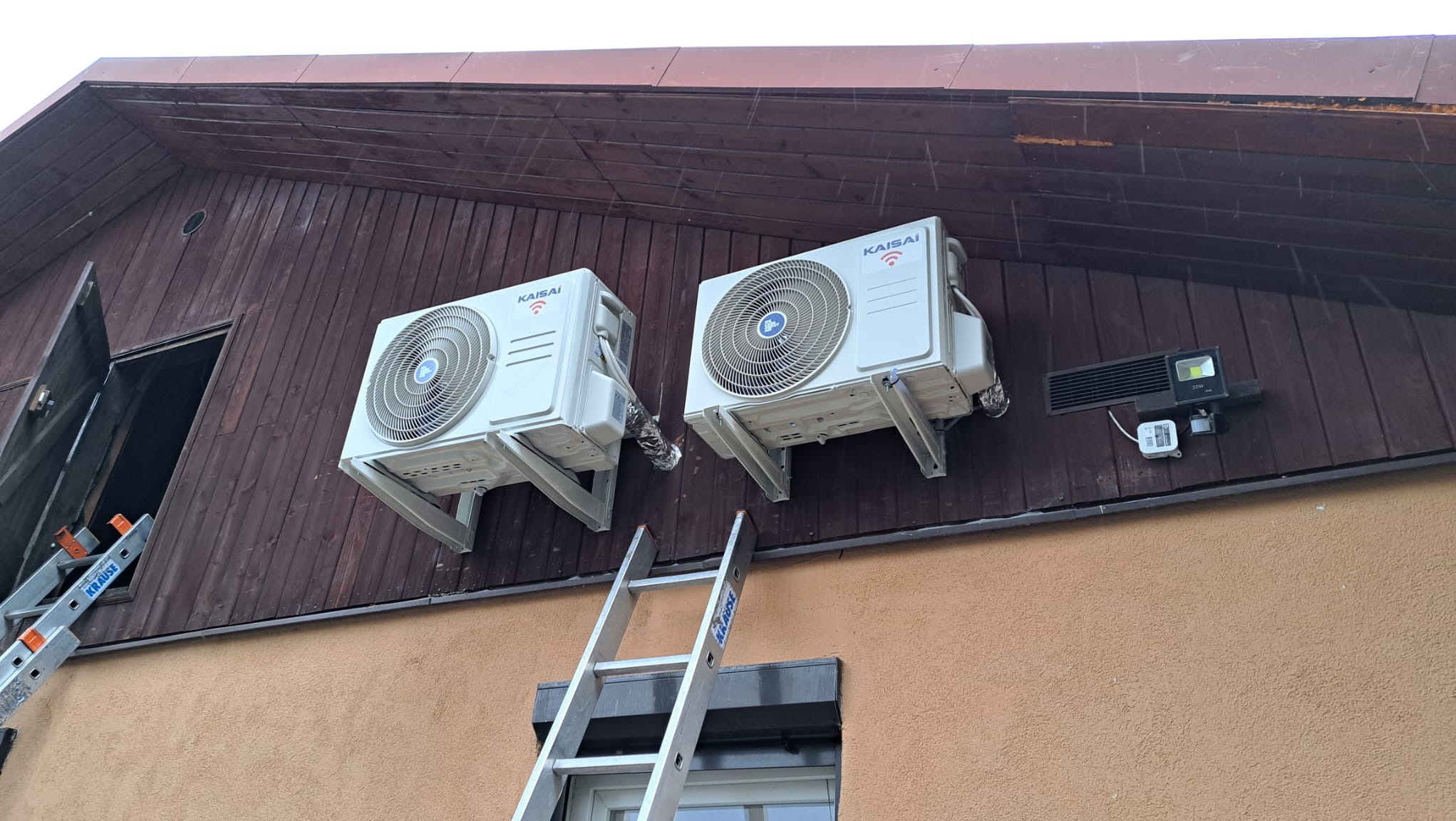 Dwa białe klimatyzatory marki Kaisai zamontowane na brązowej, drewnianej ścianie budynku, widoczne drabiny sugerujące trwający montaż, obok lampa zewnętrzna.