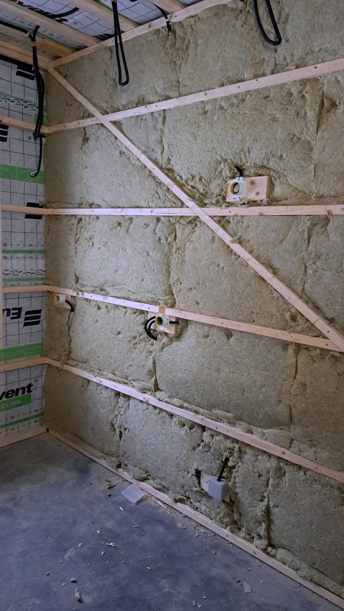 Ściana w trakcie izolacji wełną mineralną, widoczne drewniane legary, puszki elektryczne i przewody, surowy beton na podłodze.