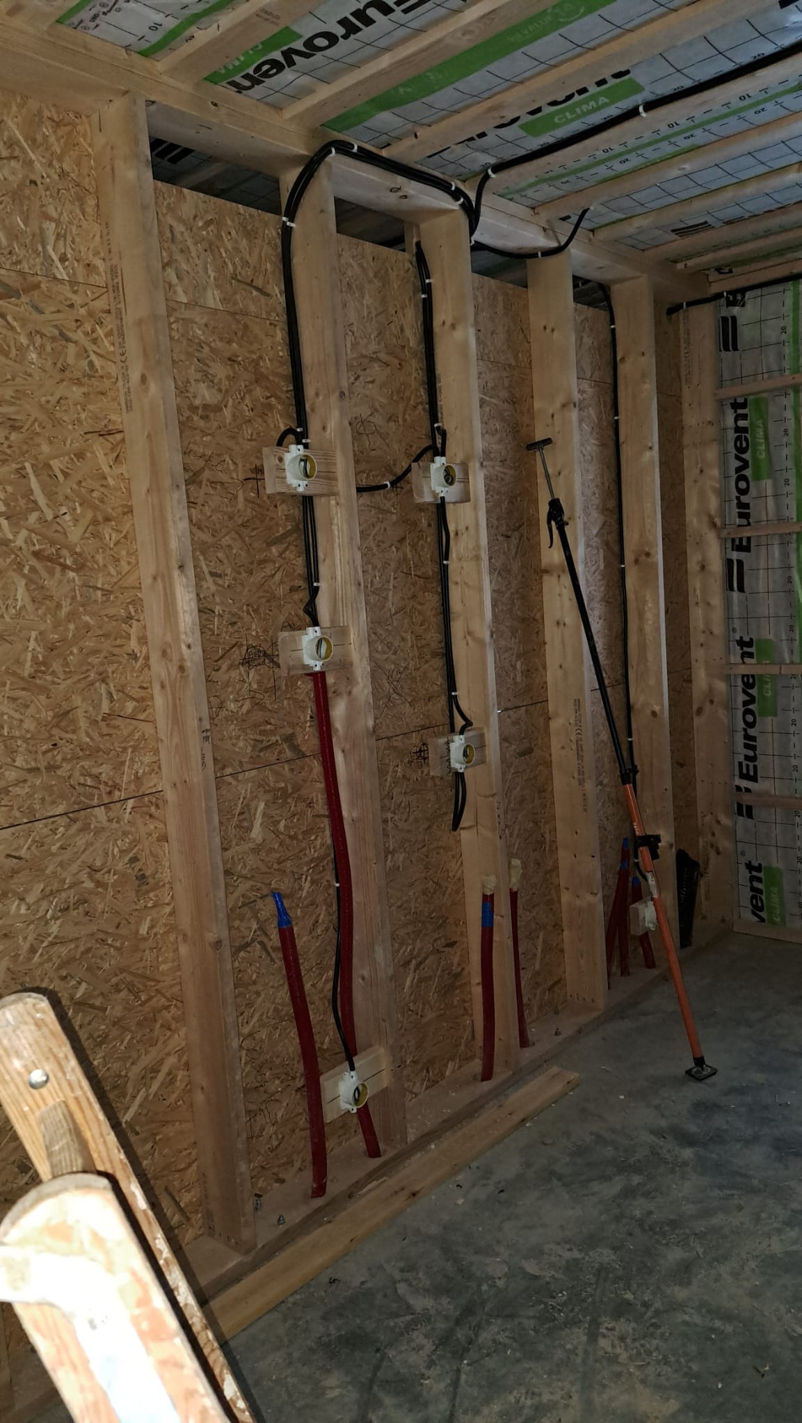 Wewnętrzna instalacja elektryczna i rury w trakcie montażu w drewnianej konstrukcji ściany, widoczne kable w peszlach i puszki elektryczne, narzędzie podpierające konstrukcję, surowy stan wykończenia.