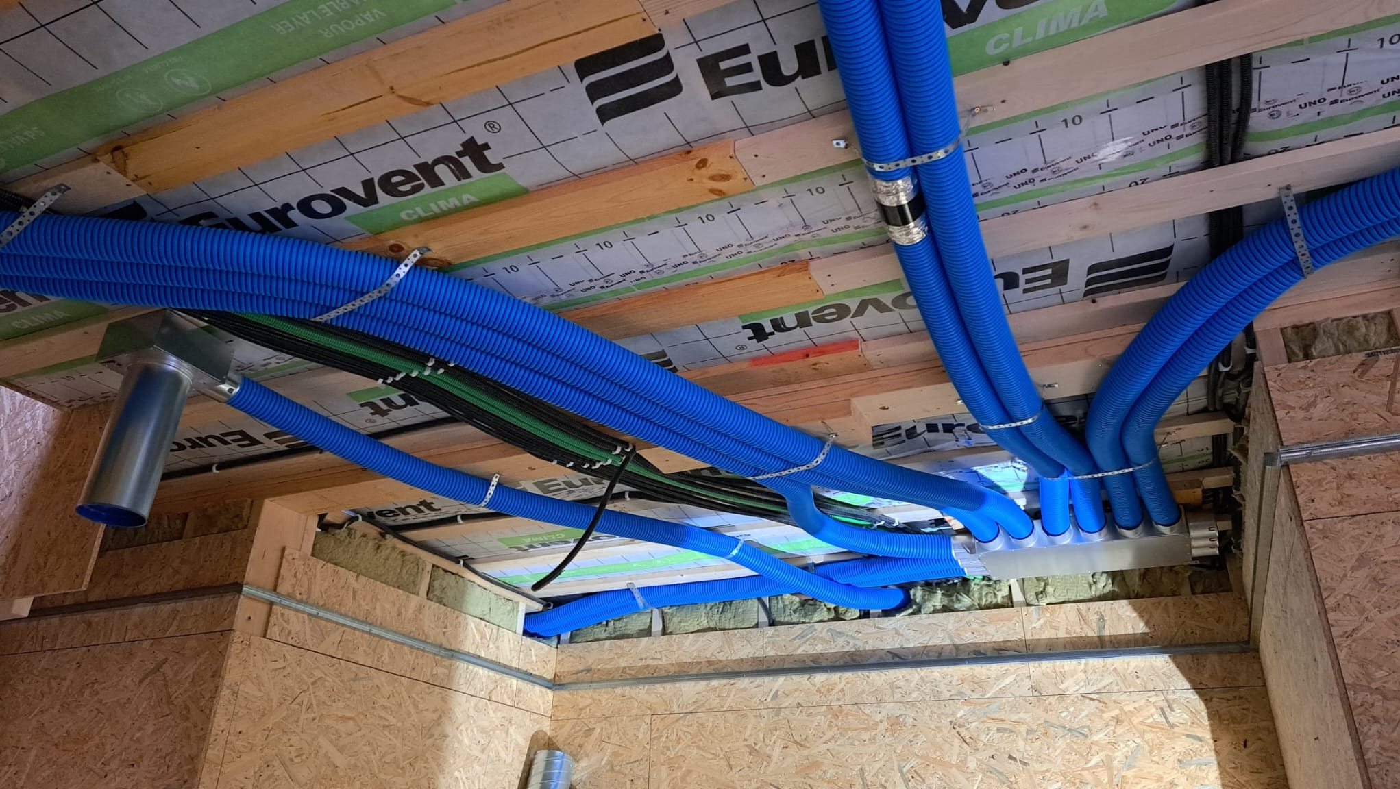 Instalacja rekuperacji z niebieskimi rurami karbowanymi, widoczne elementy montażowe, izolacja, folia paroizolacyjna Eurovent Clima i drewniana konstrukcja poddasza.