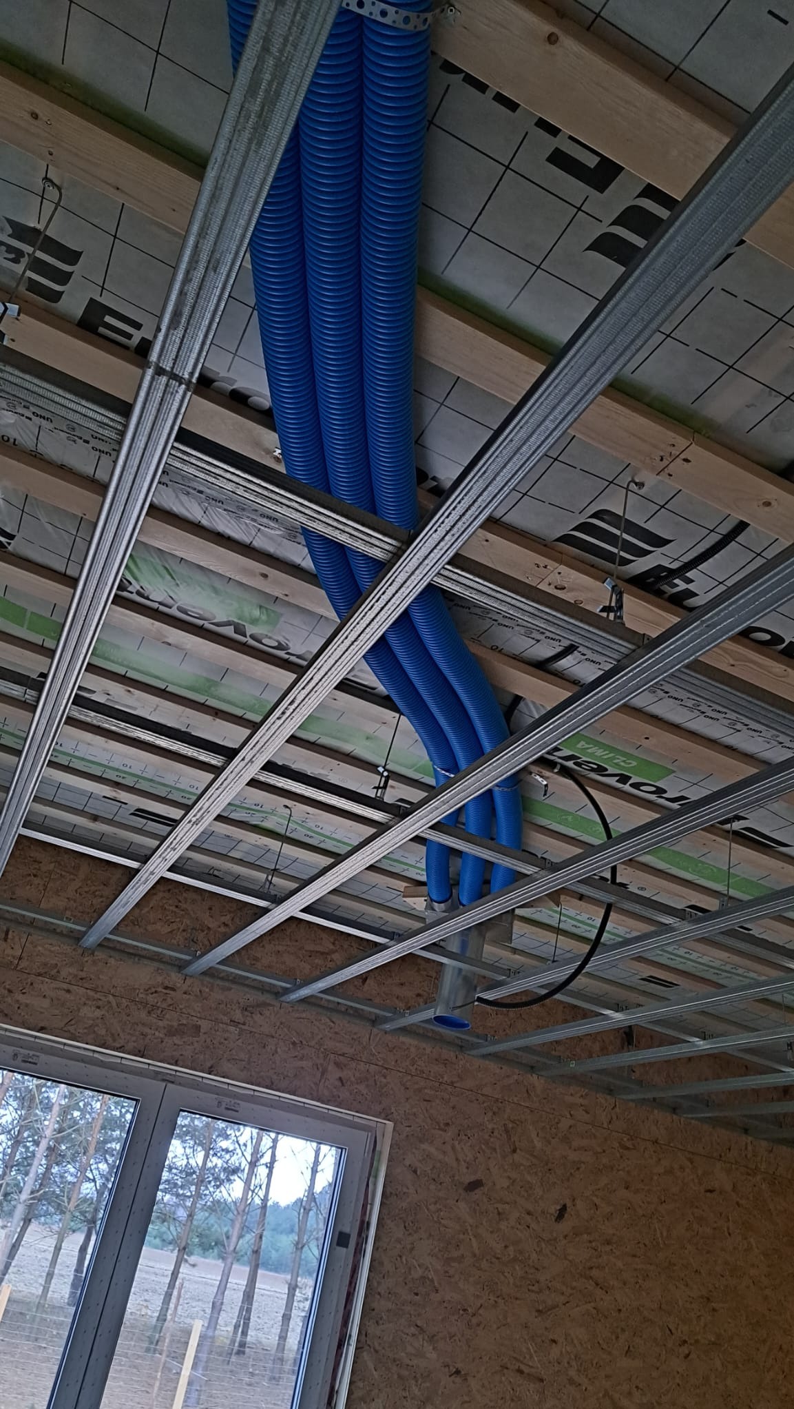 Niebieskie, karbowane rury wentylacyjne poprowadzone pod sufitem w trakcie montażu, widoczne metalowe profile i elementy konstrukcji drewnianej.