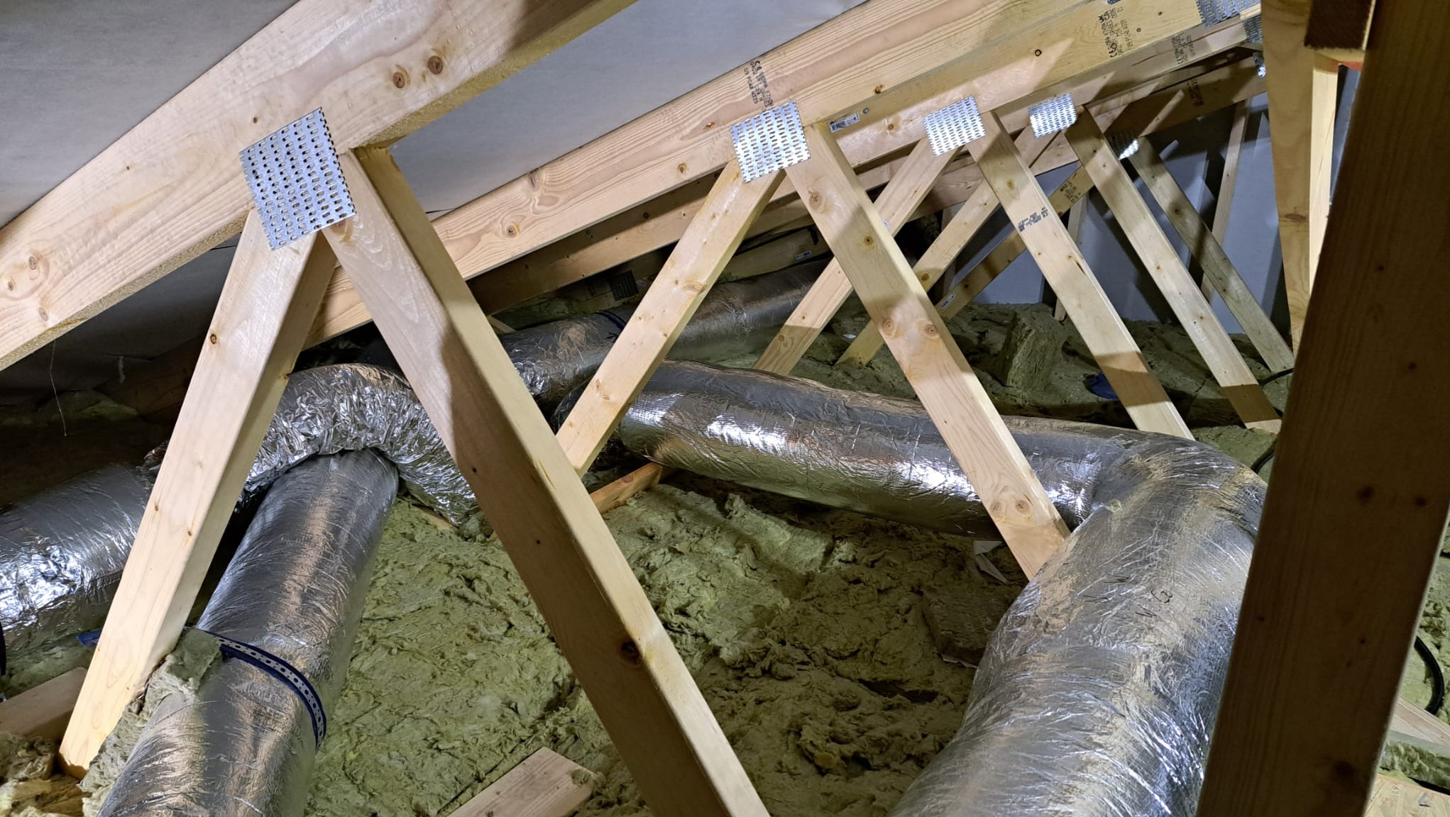 Dwa srebrne, izolowane przewody wentylacyjne umieszczone między drewnianymi krokwiami w przestrzeni poddasza, widoczne metalowe łączniki krokwi.