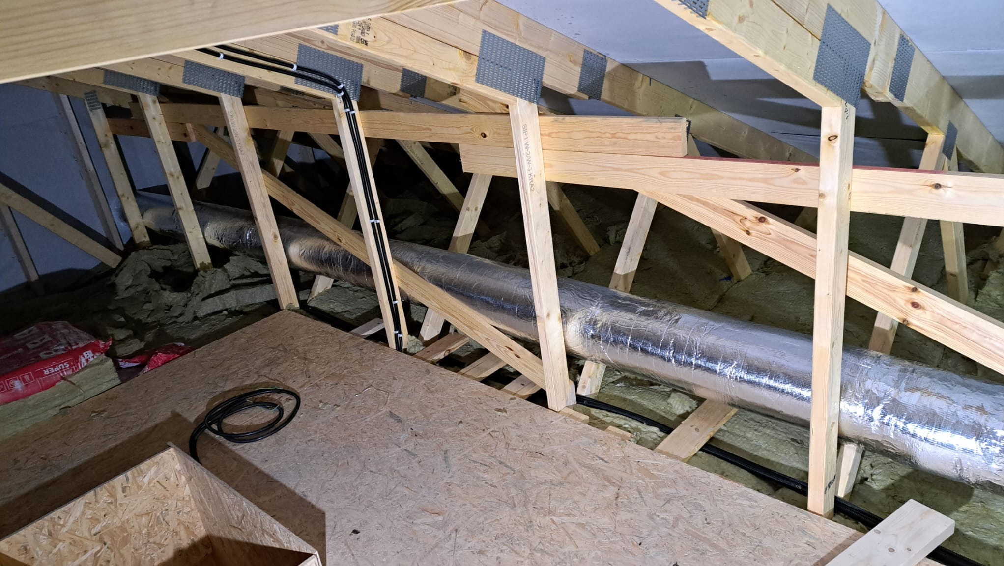 Instalacja rekuperacji w przestrzeni poddasza: widoczne rury w srebrnej izolacji, drewniana konstrukcja dachu, czarne kable elektryczne przymocowane do belek, oraz częściowo ułożona podłoga z płyt...