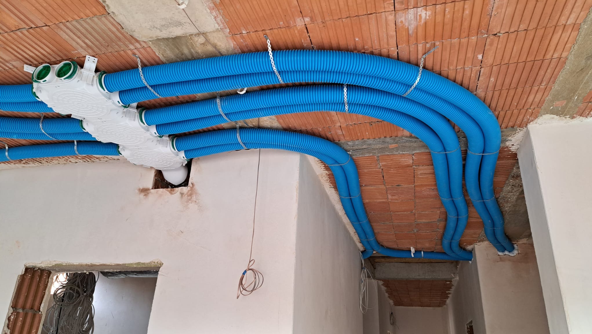 Niebieskie rury wentylacyjne zamontowane pod ceglanym sufitem, połączone z białymi rozdzielaczami, widoczne niedokończone ściany.