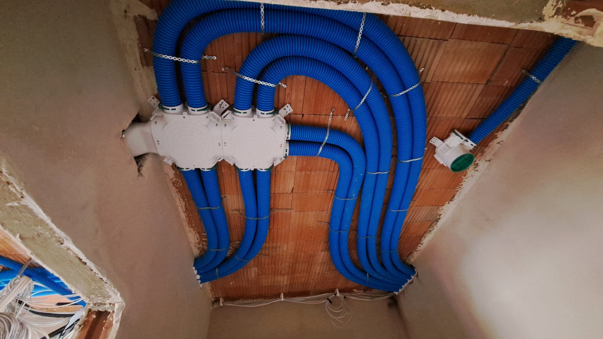 Niebieskie, karbowane rury wentylacyjne podłączone do białej skrzynki rozdzielczej na surowym, ceglanym stropie, zabezpieczone metalowymi uchwytami.