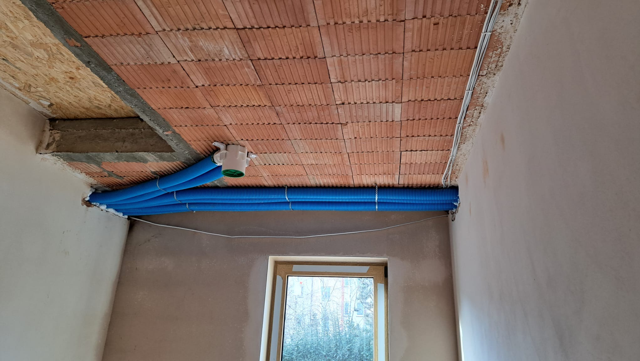 Instalacja rekuperacji z niebieskimi rurami karbowanymi pod sufitem z cegły dziurawki, widoczne przewody elektryczne i okno w surowym stanie.