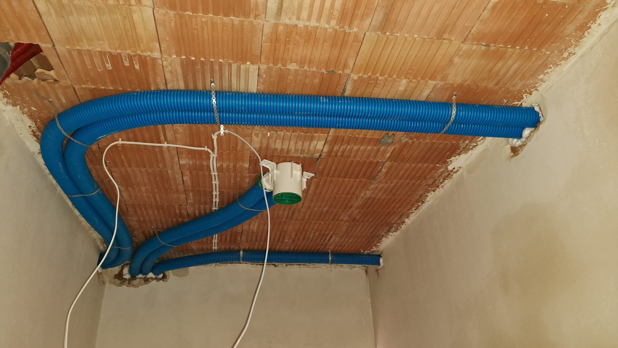 Niebieskie rury wentylacyjne przymocowane łańcuchami do sufitu z cegły w trakcie montażu, widoczne białe przewody elektryczne i fragment centrali wentylacyjnej z zielonym elementem.