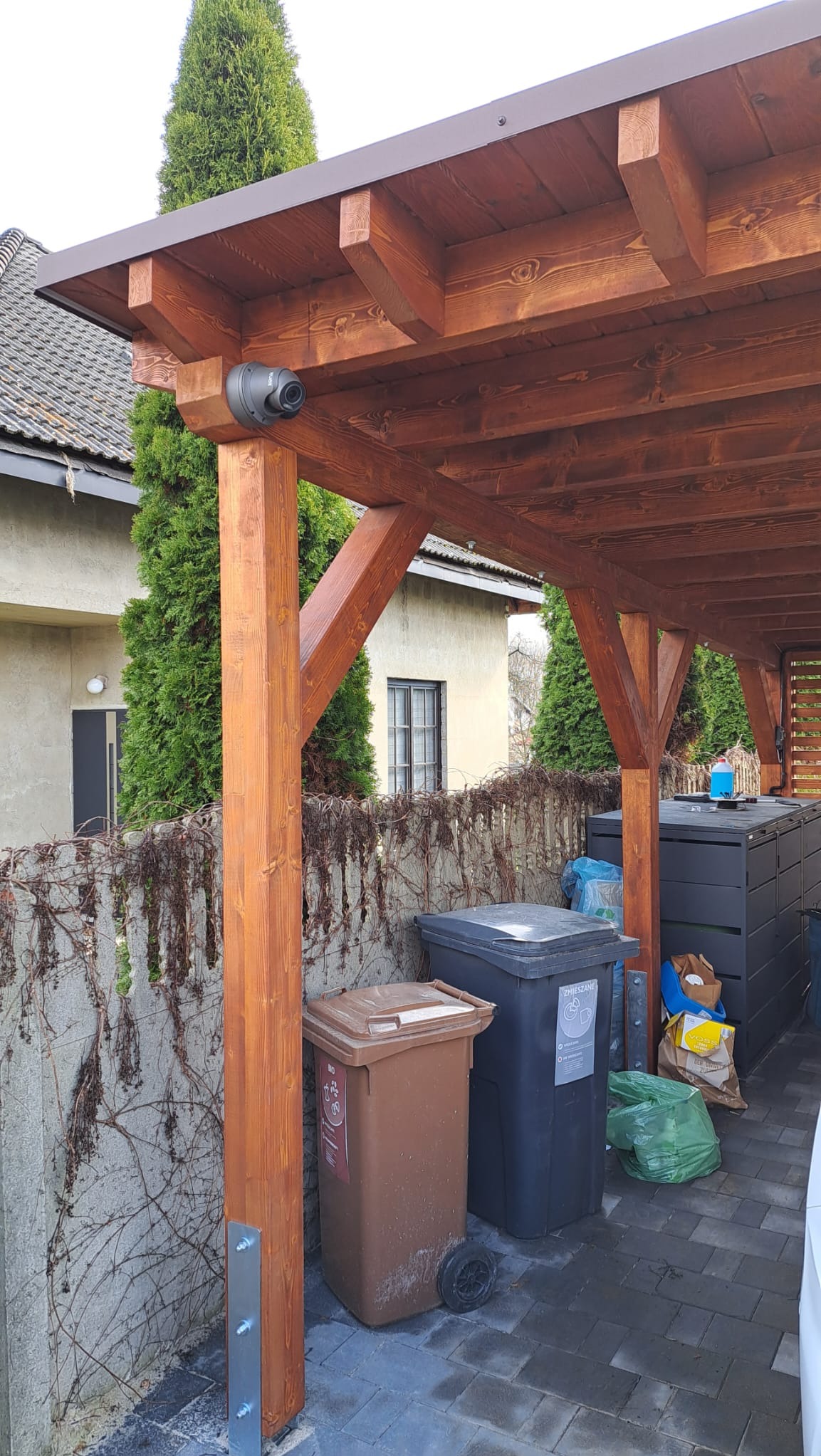 Kamera kopułkowa zamontowana pod drewnianą konstrukcją z widocznymi elementami mocującymi i śmietnikami w tle.