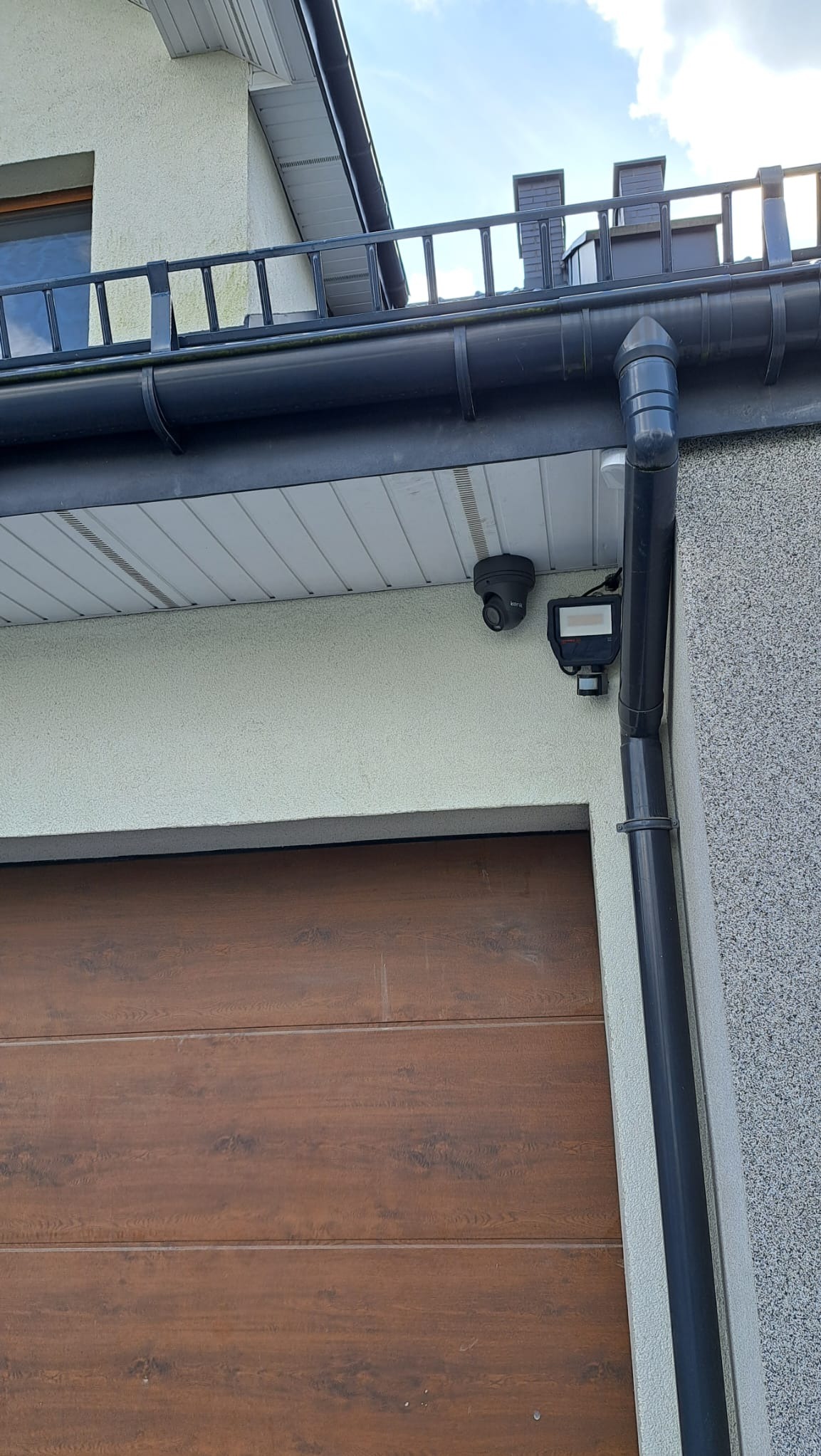 Kamera monitoringu i czujnik ruchu zamontowane nad brązową bramą garażową w Mstowie, widoczne orynnowanie i fragment elewacji budynku.