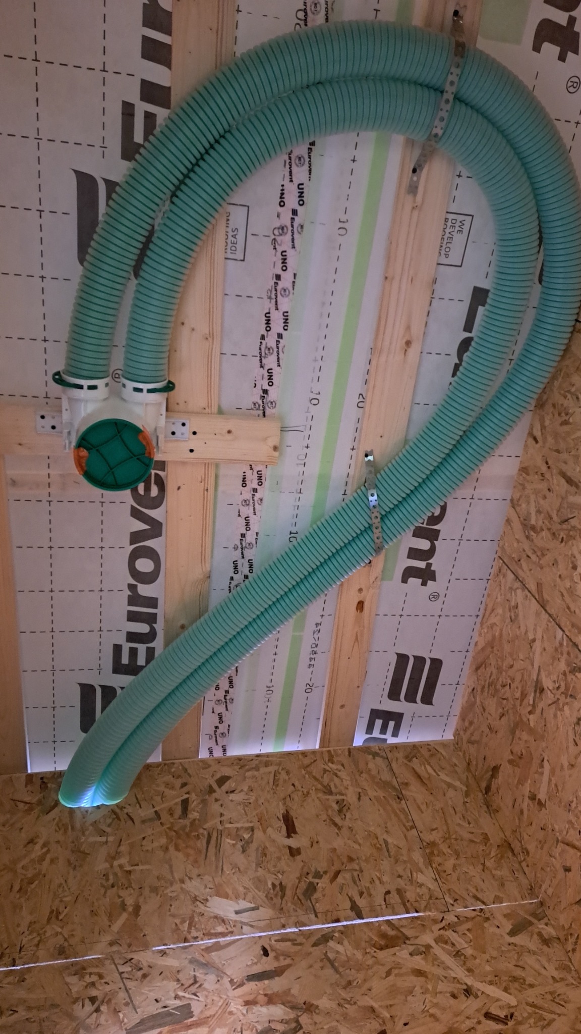 Fragment instalacji rekuperacji z dwoma elastycznymi, karbowanymi przewodami w kolorze miętowym, przymocowanymi do drewnianej konstrukcji pod stropem. Widoczne elementy mocujące i izolacja.