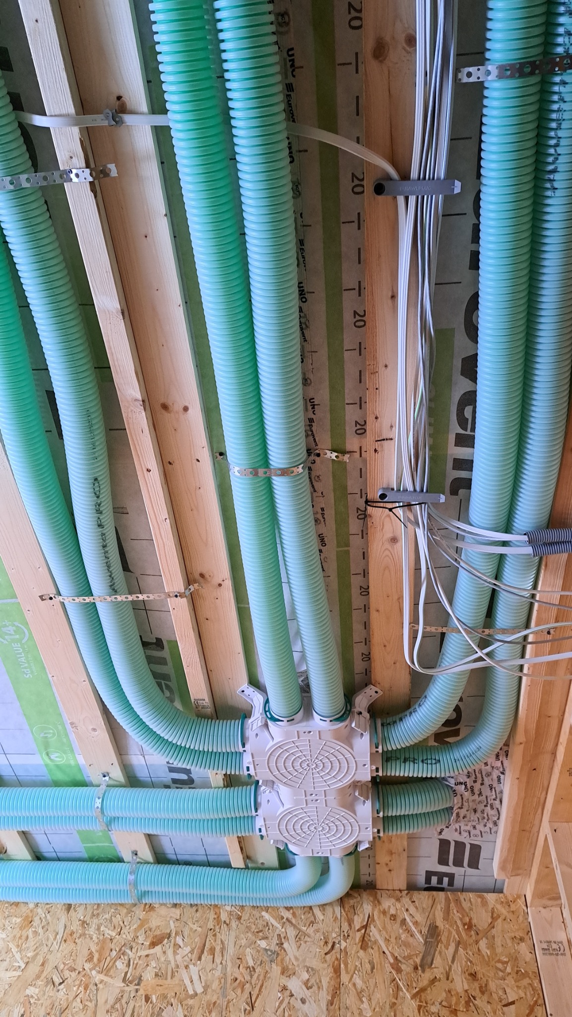 Widok od dołu na montaż systemu wentylacji mechanicznej z rurami w kolorze miętowym, przymocowanymi do drewnianej konstrukcji stropu za pomocą metalowych uchwytów i białej skrzynki rozdzielczej.