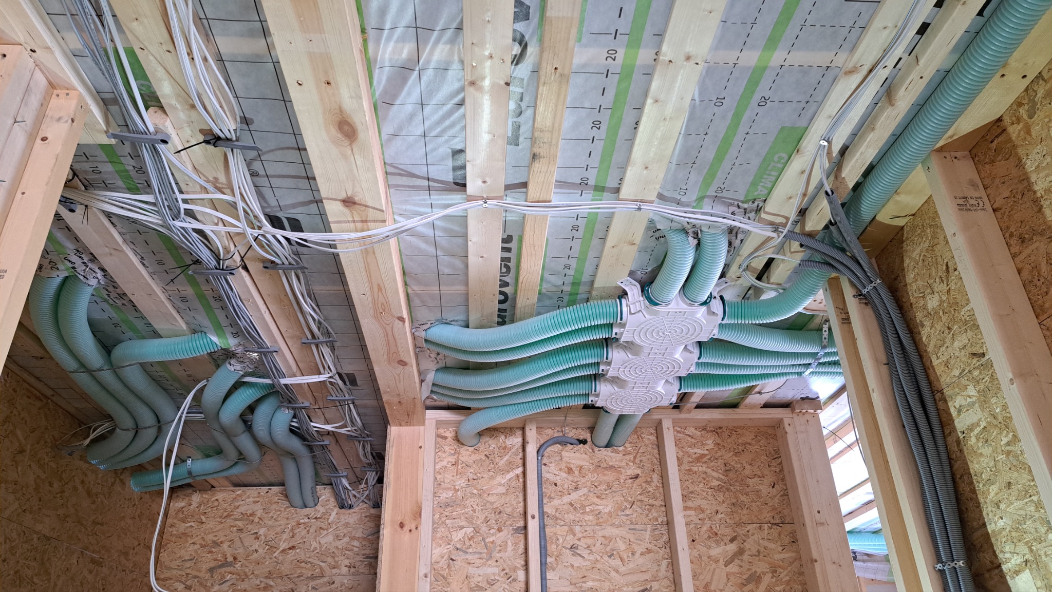 Instalacja rekuperacji i przewodów elektrycznych w surowym drewnianym wnętrzu, widoczne rury wentylacyjne w kolorze seledynowym i kable elektryczne przymocowane do drewnianej konstrukcji stropu.