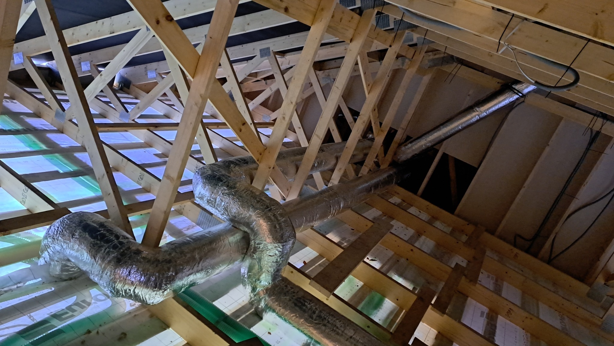 Widok z dołu na konstrukcję dachu z widocznymi rurami wentylacyjnymi w izolacji aluminiowej, zamontowanymi między drewnianymi belkami.