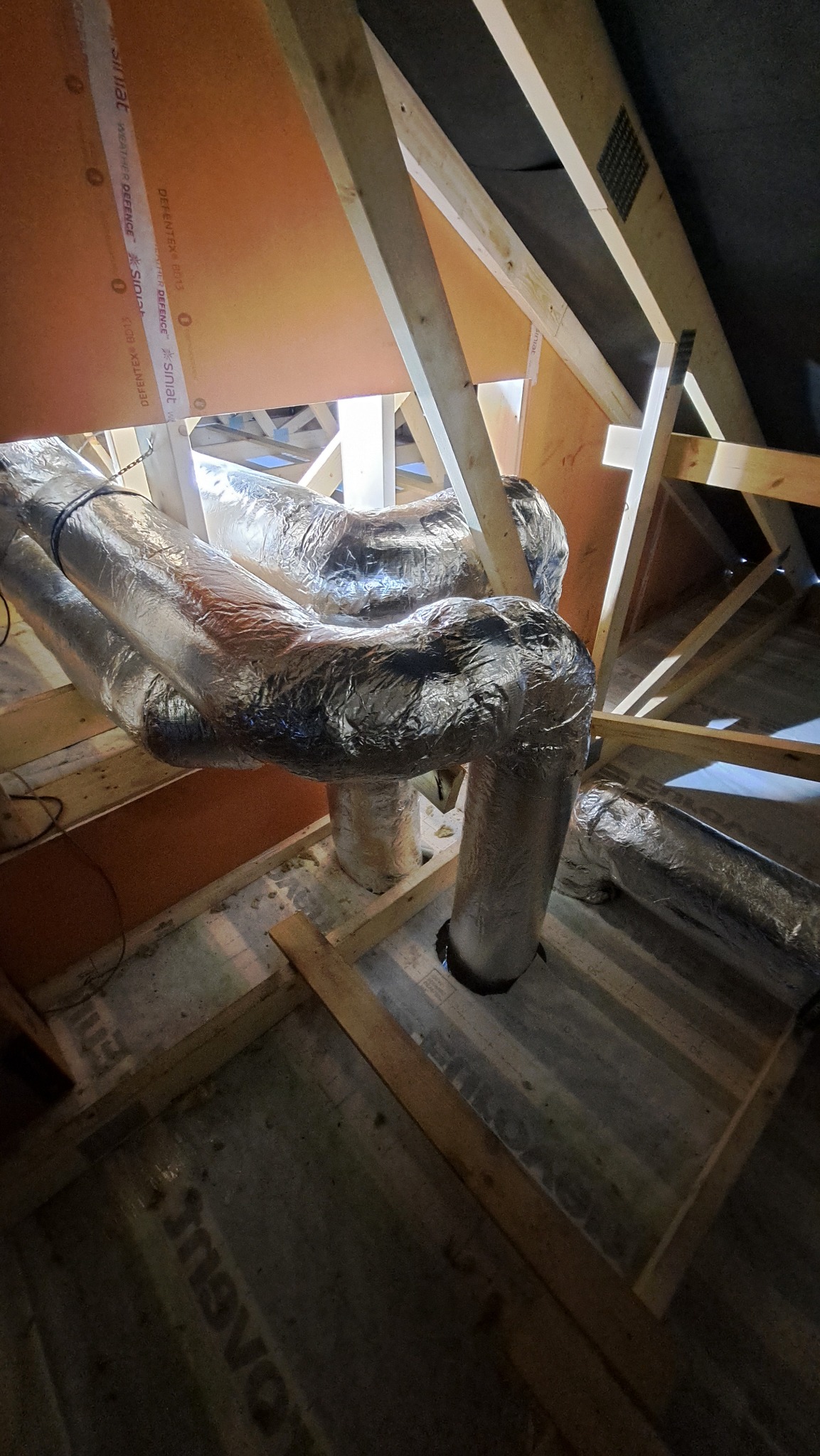 Srebrne, izolowane przewody wentylacyjne ułożone w przestrzeni poddasza z widoczną drewnianą konstrukcją dachu i deskami obiciowymi.