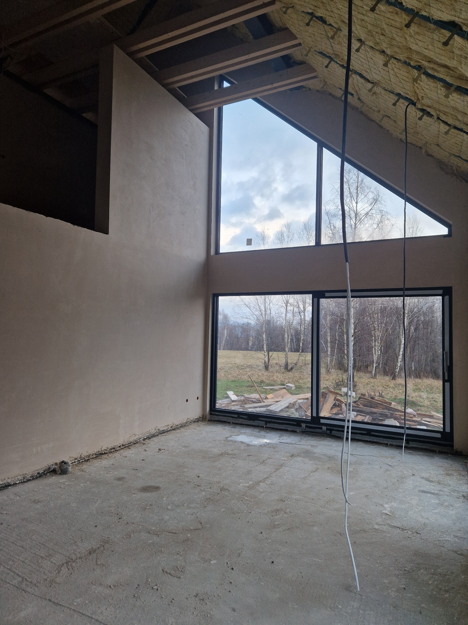 Surowe wnętrze domu w trakcie wykańczania: ściany pokryte świeżym, jasnoszarym tynkiem maszynowym, duże okna z widokiem na jesienny las, widoczne ocieplenie poddasza i kable zwisające z sufitu.
