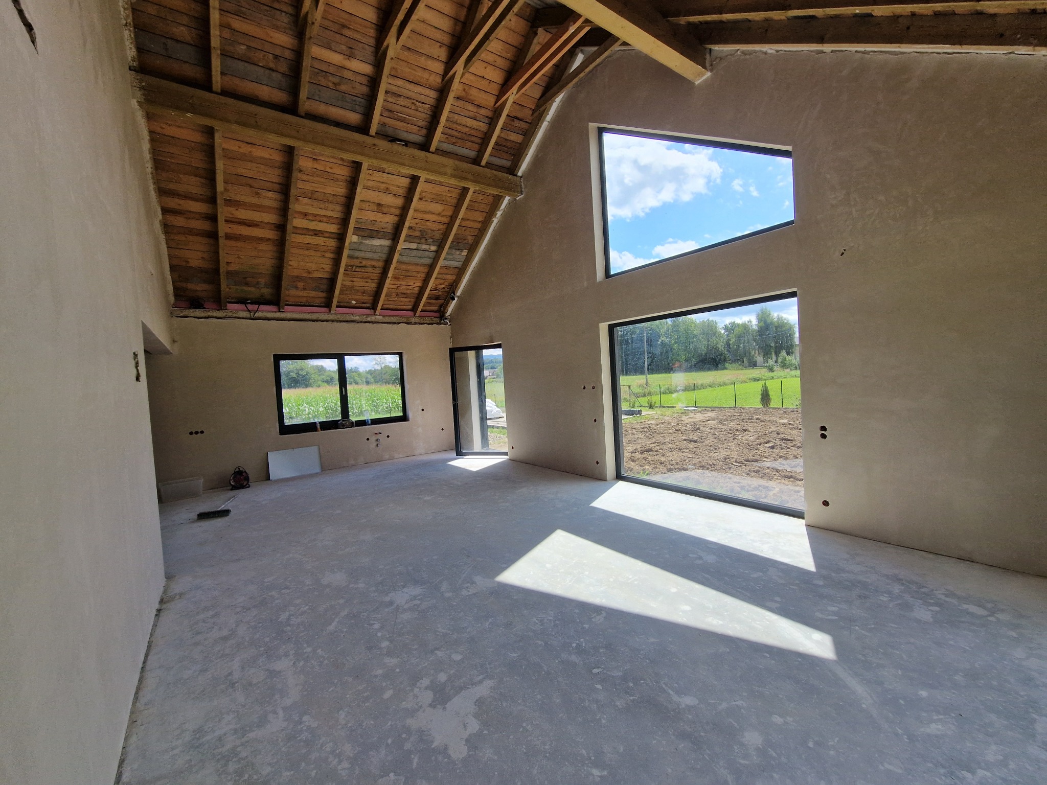 Wnętrze domu po tynkowaniu maszynowym: surowe ściany, drewniany strop, duże okna z widokiem na zewnątrz. Minimalistyczny, nowoczesny styl wykończenia.