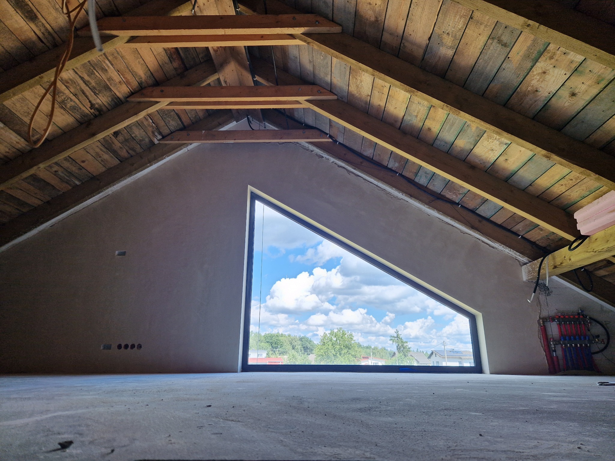 Poddasze ze świeżo otynkowanymi ścianami i trójkątnym oknem z widokiem na zewnątrz. Widoczne drewniane elementy konstrukcji dachu i instalacja grzewcza.