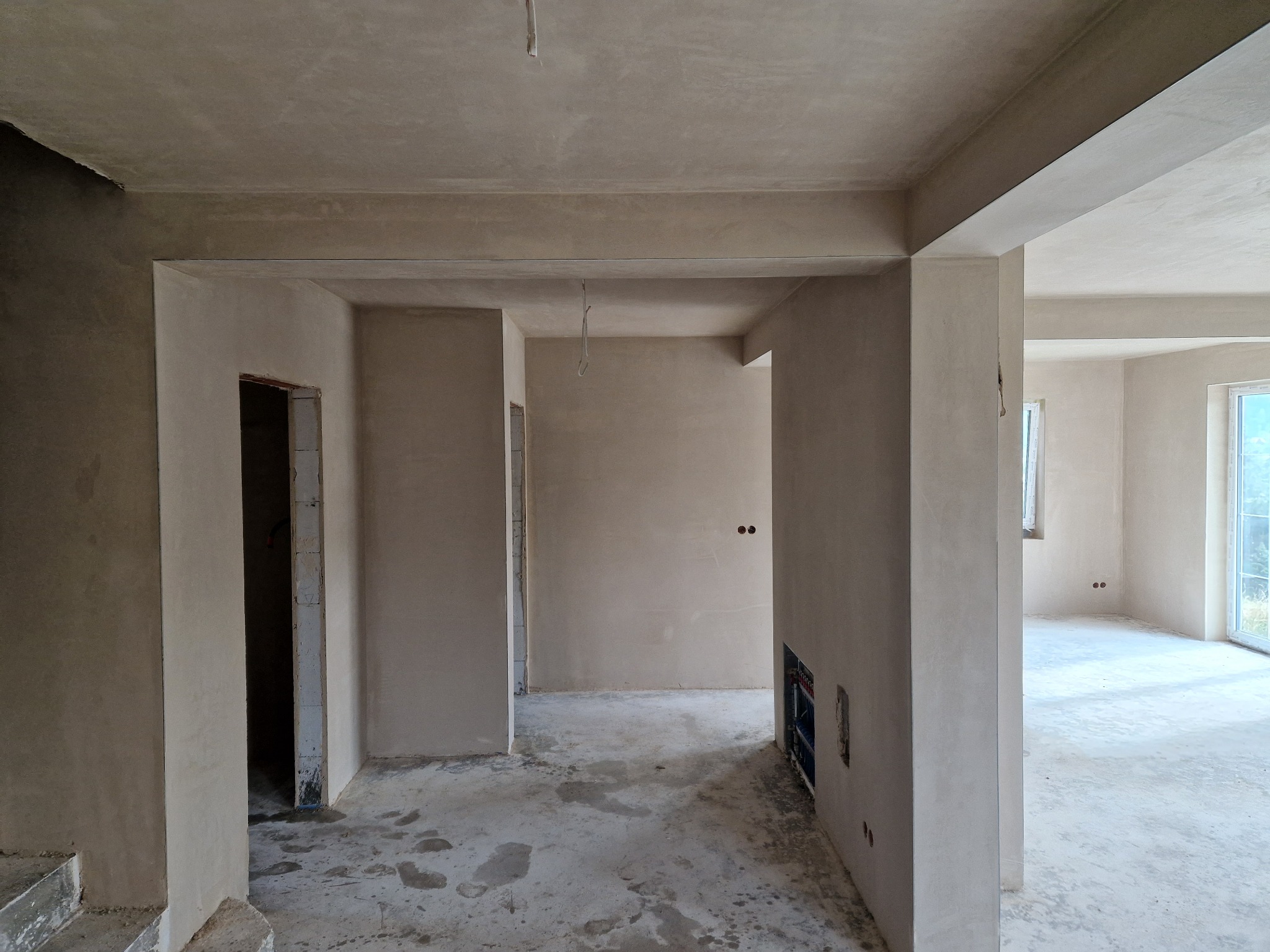 Wnętrze domu w trakcie prac wykończeniowych, ściany pokryte świeżym tynkiem, widoczne instalacje elektryczne i hydrauliczne w ścianach, surowy beton na podłodze.