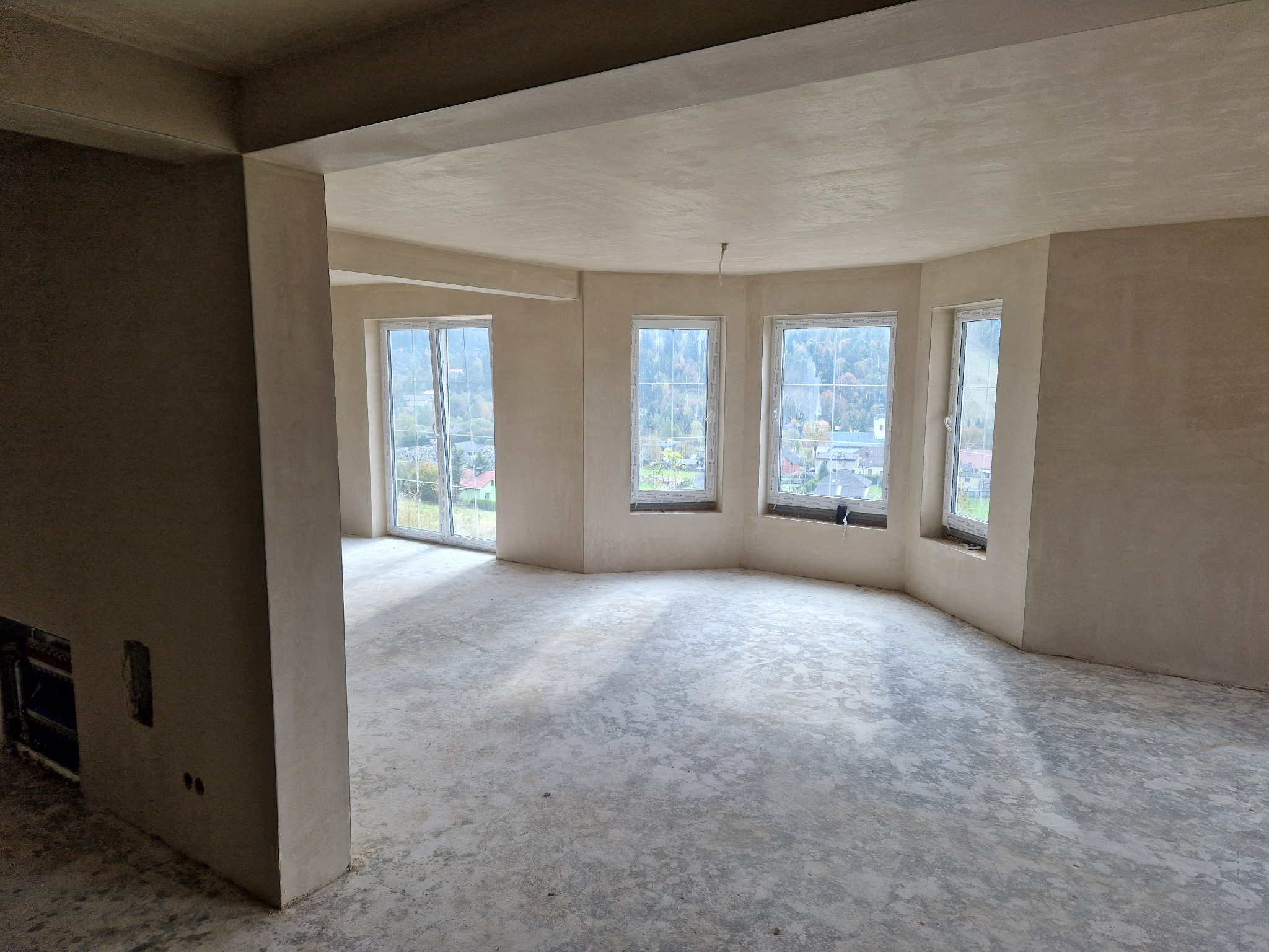 Surowe wnętrze po nałożeniu tynków, widoczne okna z widokiem na zieleń, surowy beton na podłodze, przygotowane do dalszych prac wykończeniowych.