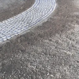 Zakrzywiona ścieżka z jasnoszarej kostki granitowej prowadzi przez świeżo przygotowany teren w kierunku metalowej bramy w tle, widoczny wąż ogrodowy.