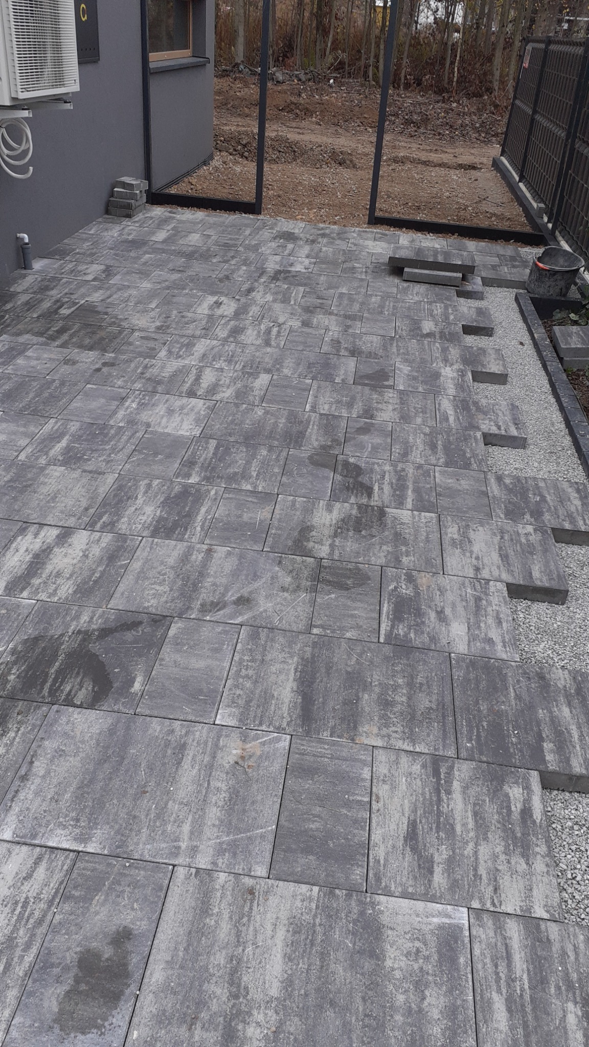 Fragment układanej nawierzchni z szarych, prostokątnych płyt betonowych o nieregularnej fakturze, widoczne przygotowane podłoże z drobnego żwiru i niedokończone obrzeże, w tle szara elewacja...
